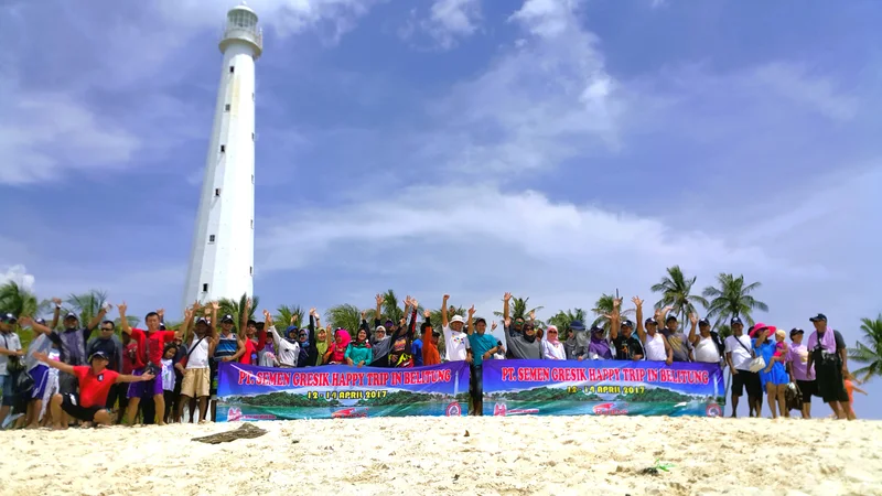 Grup perjalanan Ruhens dengan spanduk di pantai Belitung, dengan mercusuar di belakangnya.