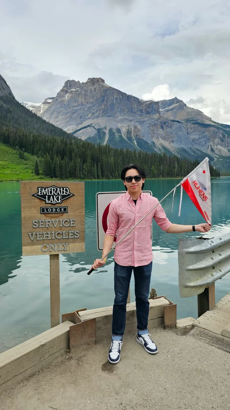 Seorang peserta memegang bendera Ruhens di tepi Emerald Lake dengan hutan dan pegunungan di belakangnya.