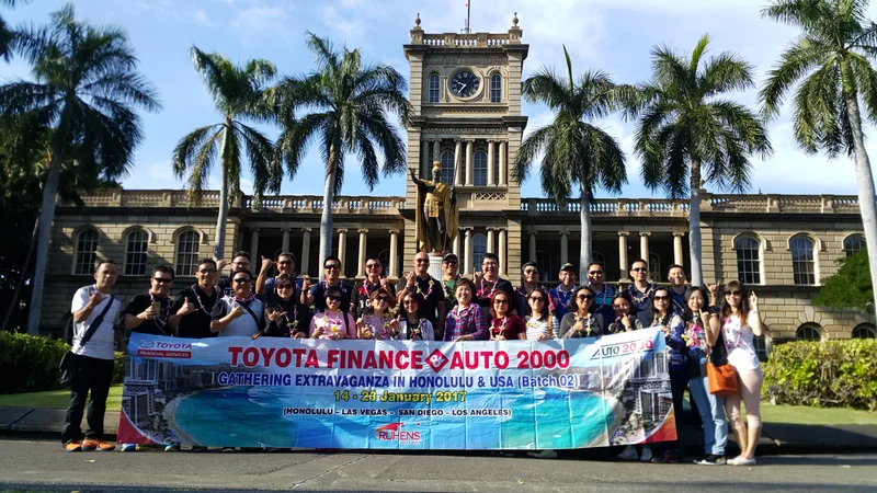 Grup perjalanan Ruhens dengan spanduk di depan bangunan bersejarah dan pohon palem di Hawaii.
