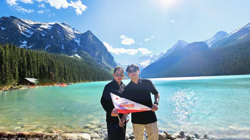 Dua peserta memegang bendera Ruhens di tepi Lake Louise dengan danau dan pegunungan di belakangnya.