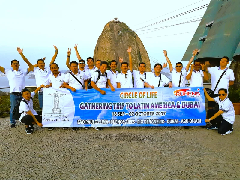 Grup perjalanan Ruhens dengan spanduk di depan gunung dan kereta gantung di Rio de Janeiro.