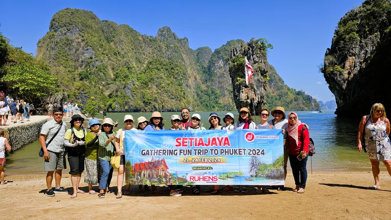 Grup perjalanan dengan spanduk Setiajaya berfoto di pantai dengan tebing batu di belakangnya.