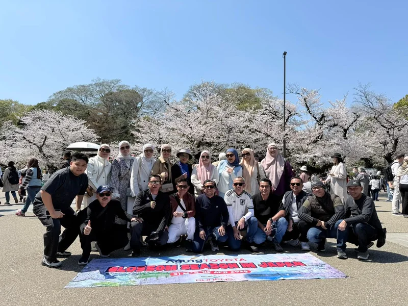 Grup perjalanan dengan spanduk berfoto di depan pohon sakura yang sedang berbunga.