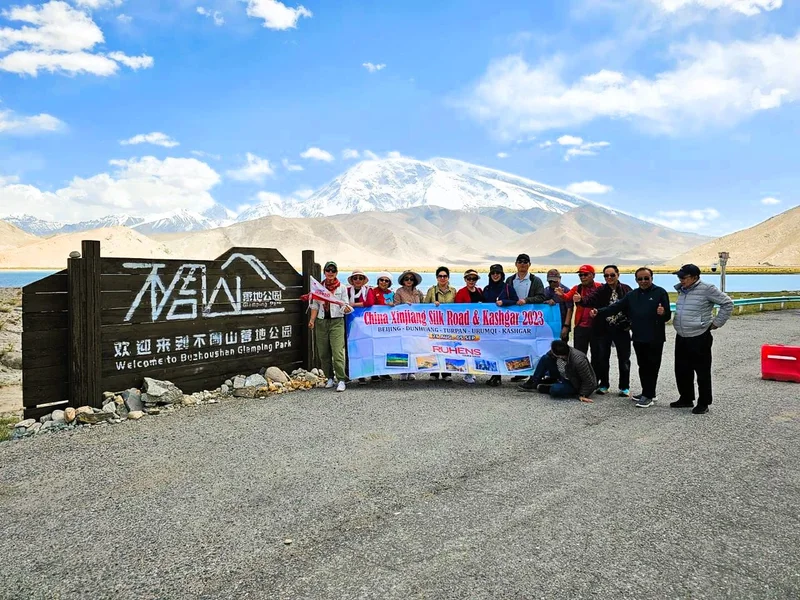 Grup perjalanan dengan spanduk China Xinjiang Silk Road berfoto di depan papan taman dan pegunungan bersalju.