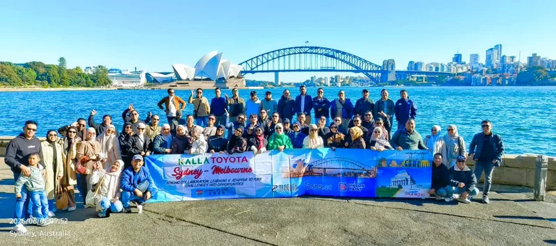 Grup perjalanan dengan spanduk Kalla Toyota berfoto di tepi pelabuhan Sydney dengan gedung opera dan jembatan di belakangnya.